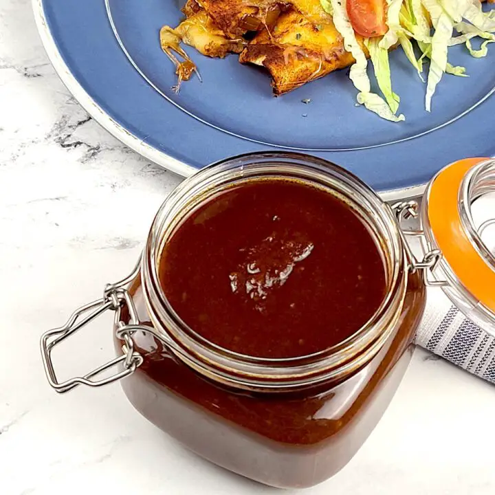 Homemade Red Enchilada Sauce in a lidded jar. In the background is a blue plate with cheese enchiladas topped with shredded lettuce.