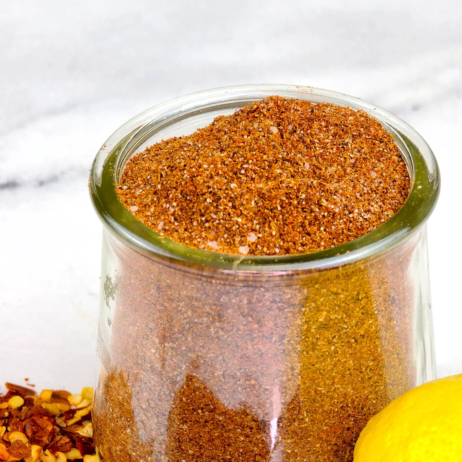 Homemade taco seasoning mix recipe in a glass jar. In the background are red pepper flakes and a lemon on a white marble background