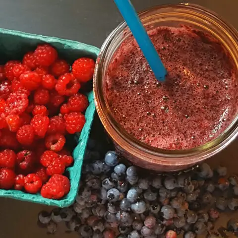 Maine berry smoothie with straw sticking out next to a box of red raspberries and bluberries on a black background