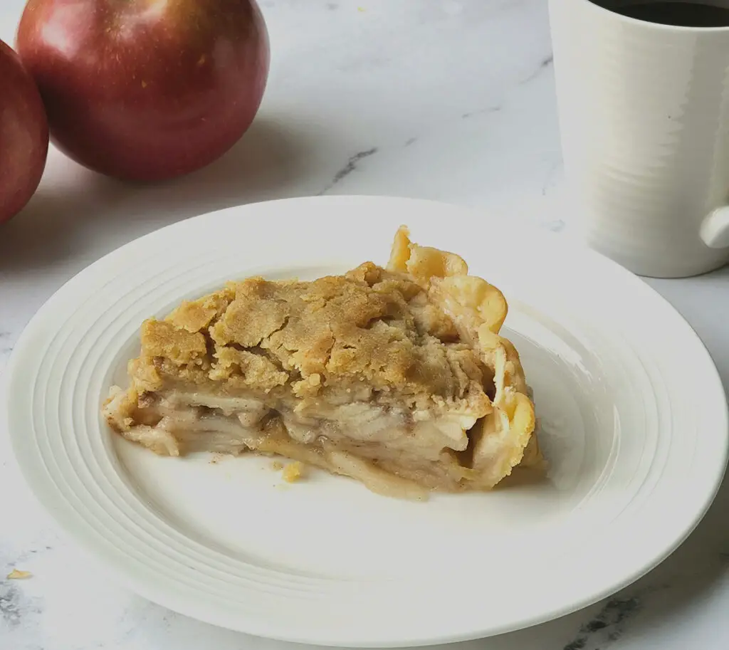 A slice of French Apple Pie on a white plate. Apples and a mug of coffee in the background. All placed on a marble surface.