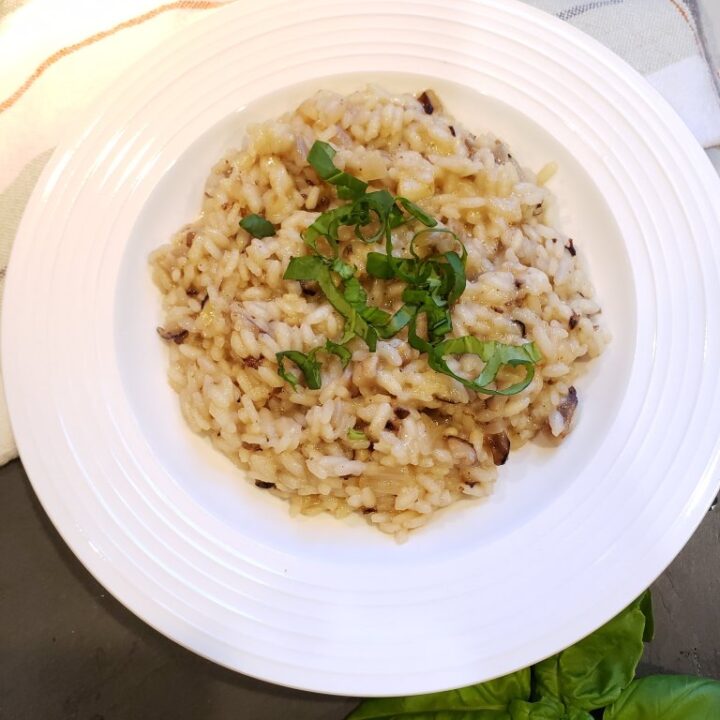 mushroom risotto recipe with basil on top in a white bowl. Bowl is on top of a back background, towel and next to basil leaves