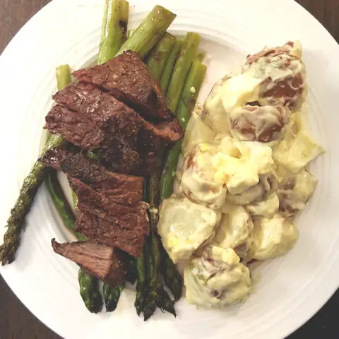steak on a bed of grilled asparagus next to potato salad. All on a white plate