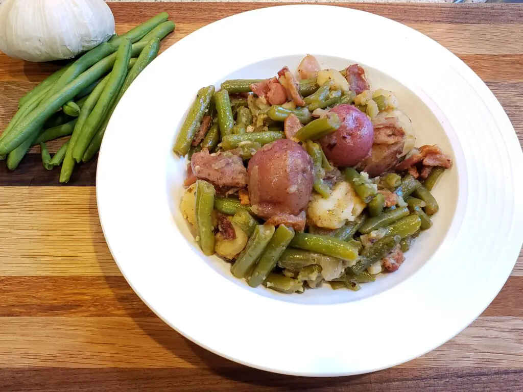 Green Beans with Red Potatoes and Bacon in a white bowl with green beans and garlic on the side