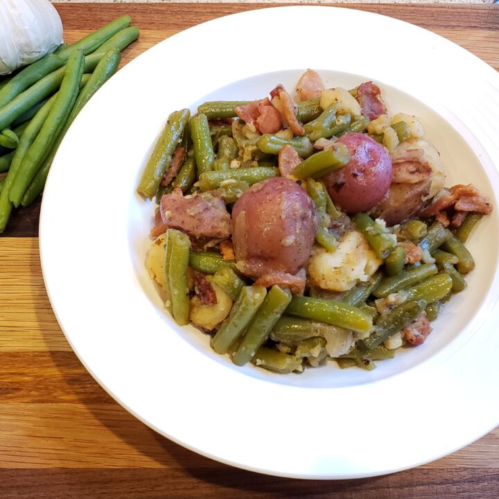 Green beans with red potatoes and bacon in a white bowl. Green beans and garlic in the corner