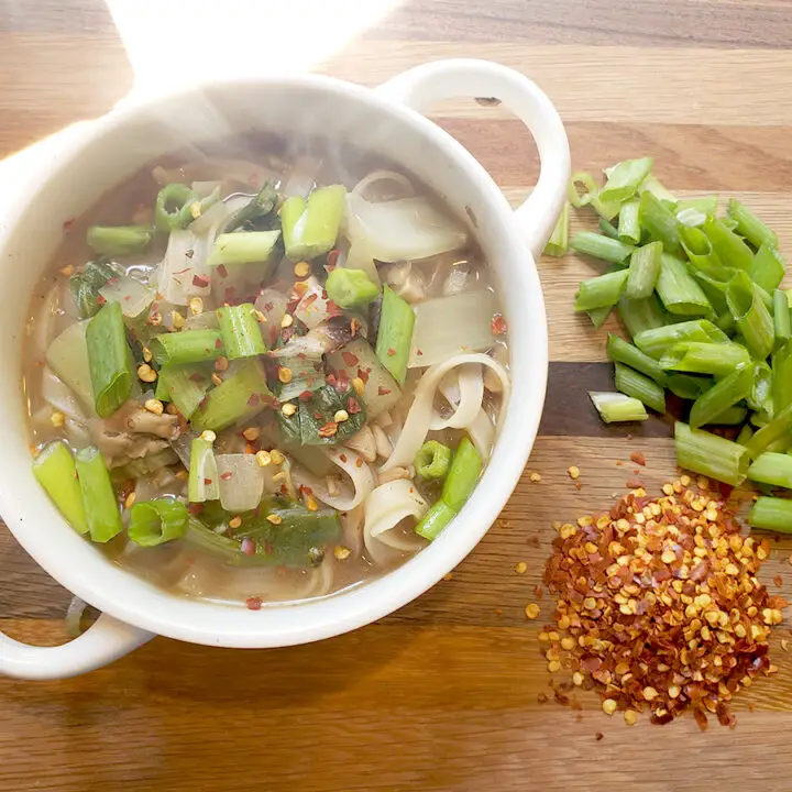 Steaming bok choy noodles in a white bowl with red pepper flakes and green onion tops sitting next to bowl on a cutting board