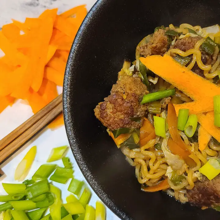 Ginger Meatball Ramen Recipe in a black bowl next to a pile of carrot shavings, pair of chopsticks, and diced green onion tops