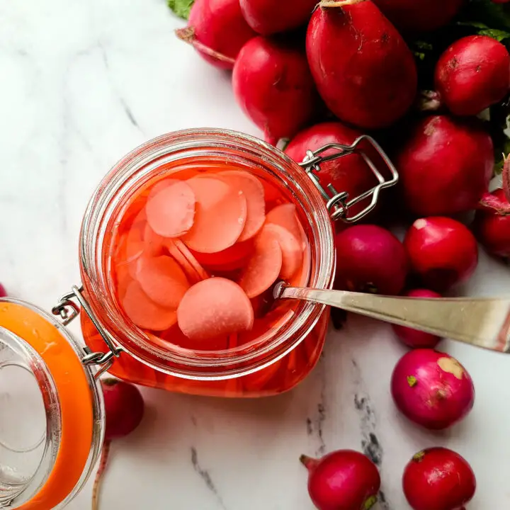 pickled radish recipe in a glass jar with a fork sticking out next to a bunch of radishes and loose radishes on a white marble background