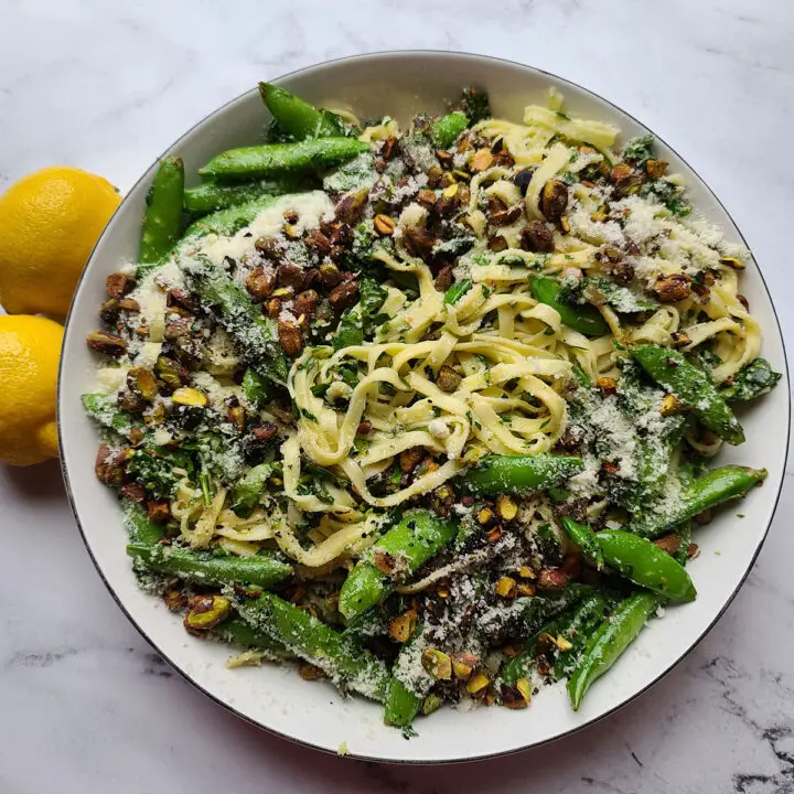 Spinach Pesto and Sugar Snap Pea Linguine topped with pistachios and parmesan cheese in a bowl next to two lemons