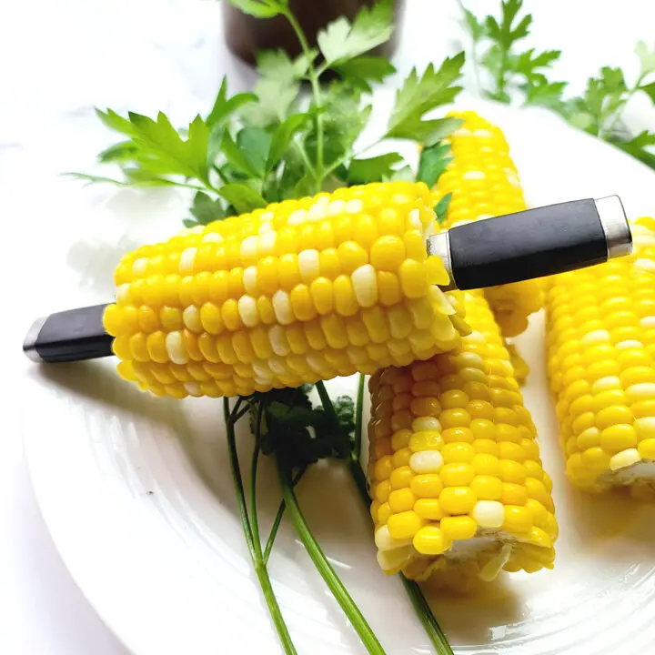 Instant Pot Corn on the cob stacked on a white plate with springs of parsley next to it and corn cob holders in the ends of one ear.