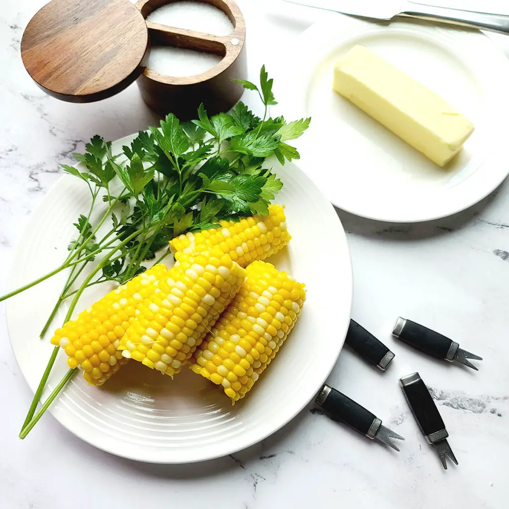 4 small ears of instant pot corn on the cob on a white plate next to several springs of parsley.  Above the plate is a brown salt keeper with salt in it, then a white plate with a stick of butter on it, then 4 corn on the cob holders. 