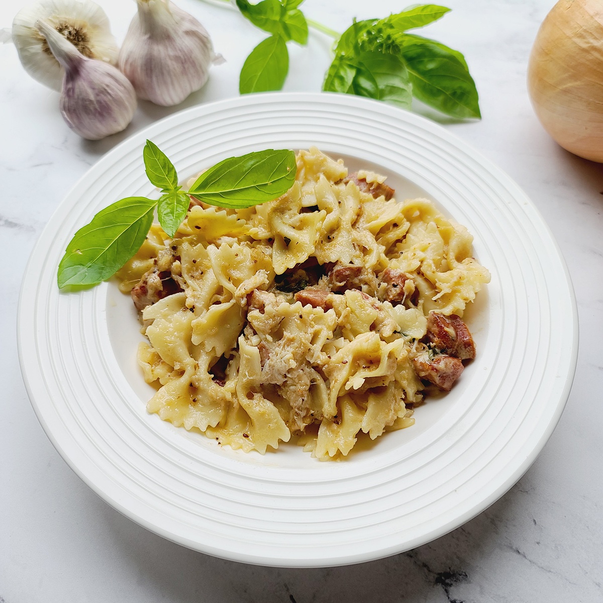 Andouille Crab Bow Tie Pasta Alfredo recipe in a white bowl with a sprig of basil in the pasta. The bowl is surrounded by garlic, a stem of bail and an onion all on a white marble backdrop