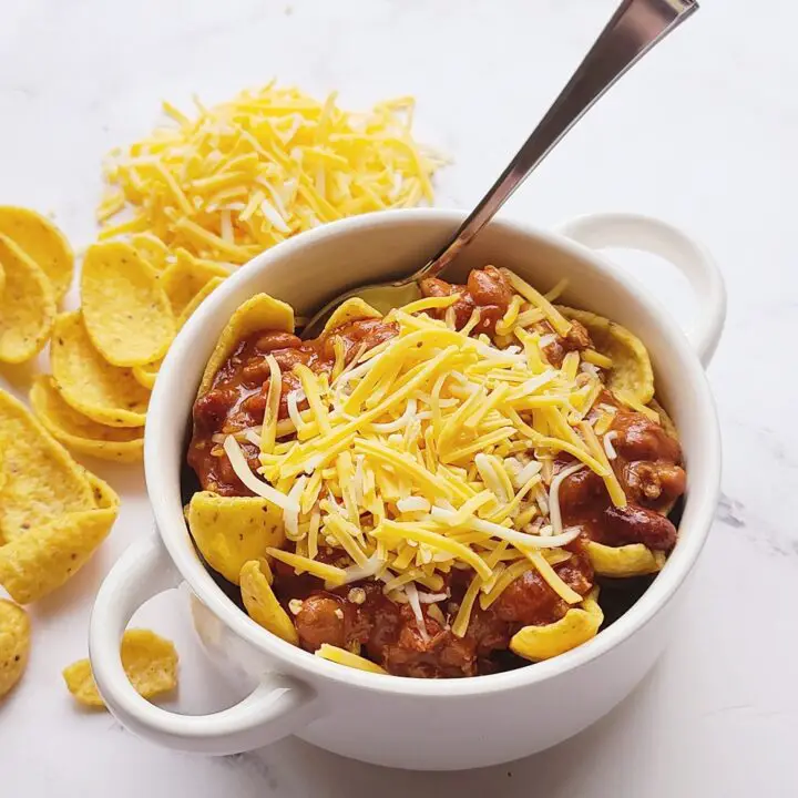 Frito Chili Pie in a white bowl with a spoon sticking out of bowl. Next to the bowl are fritos scattered on a white marble backdrop next to a pile of shredded cheese