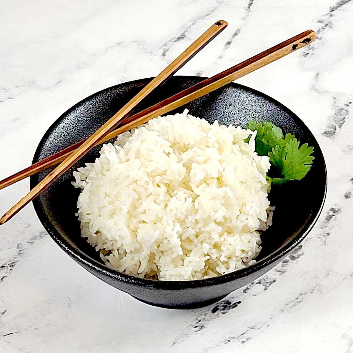 Jasmine Rice in a black bowl with cilantro leaves on the side and wood chopsticks resting on the bowl. All on a marble background