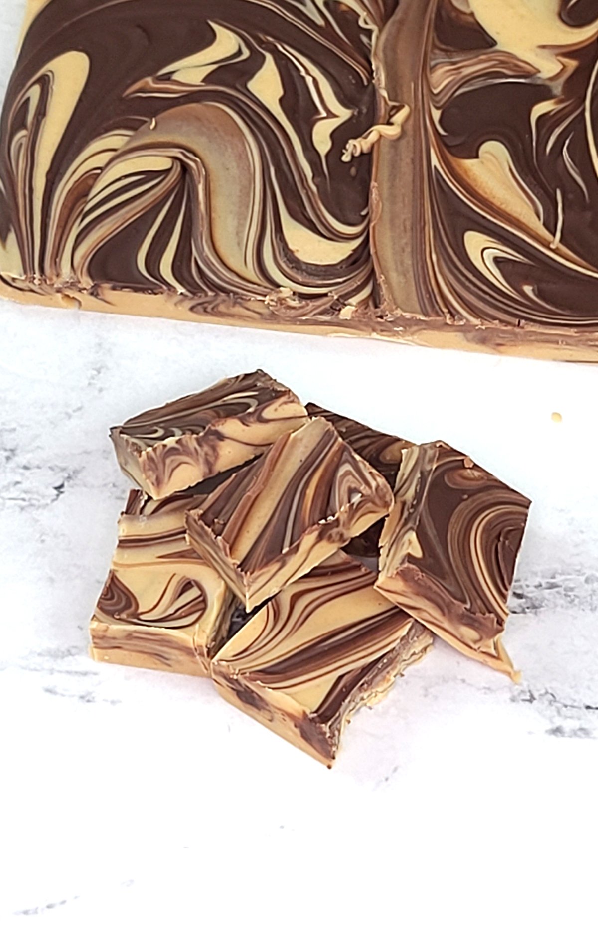 Squares of Tiger Butter Recipe piled on a white marble background. A slab of tiger butter fudge in the background