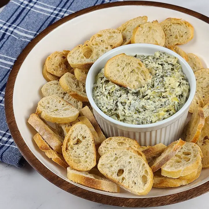 Instant Pot Spinach Artichoke dip recipe in a smaller white bowl. Surrounded by crostini in a larger white bowl. In background is a blue and white towel on a marble surface.