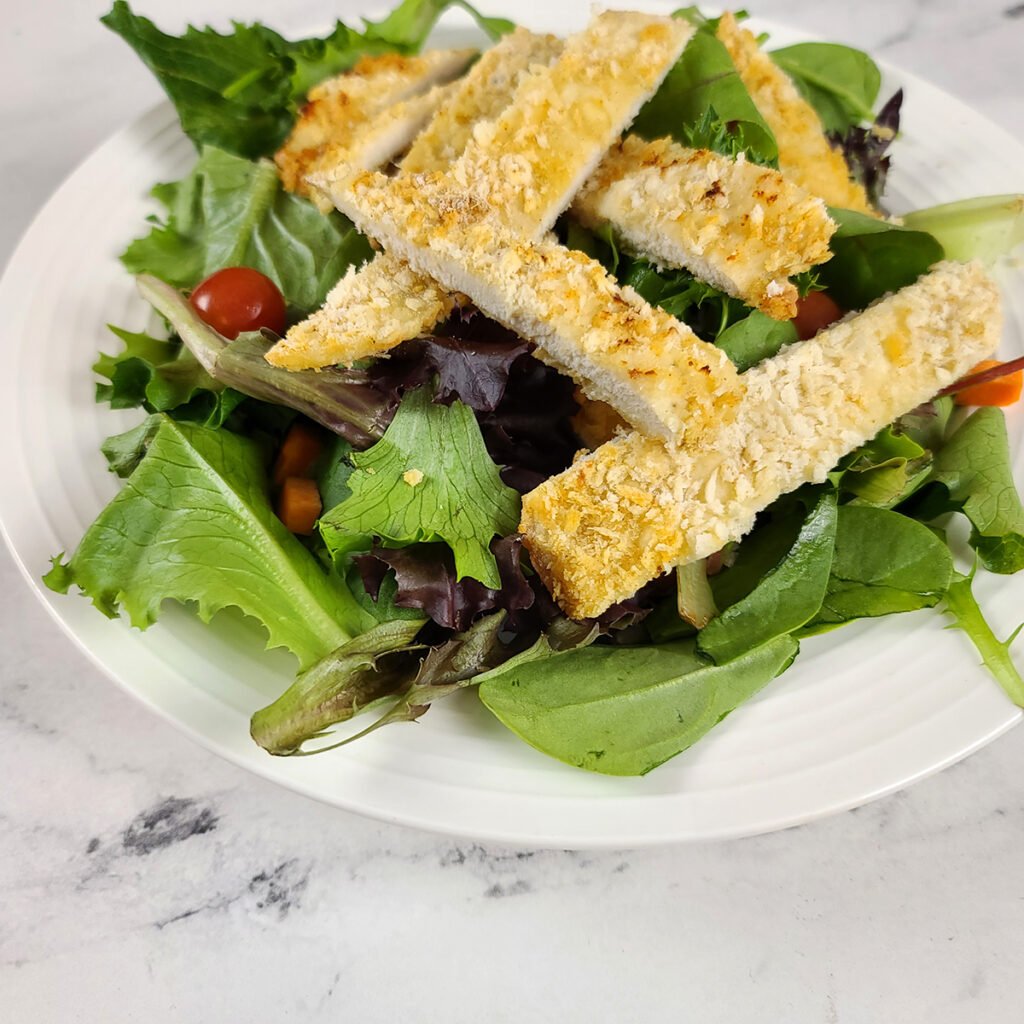 Air Fryer Chicken Cutlet cut in strips on top of a mixed greens salad with tomatoes.  In a white bowl and on a marble surfacte.